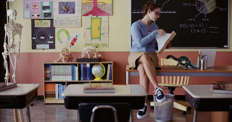 Teacher sitting on desk, reviewing collected papers in a folder inside the classroom.