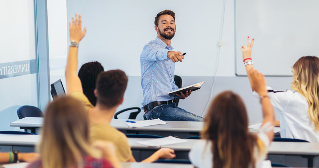 Teacher at whiteboard while students raise hands in classroom