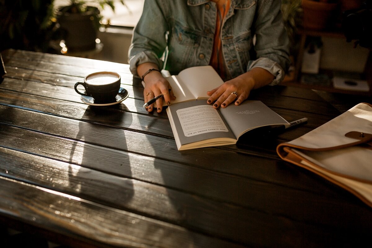 Lady sitting at a wooden table, drinking coffee, and writing in a journal