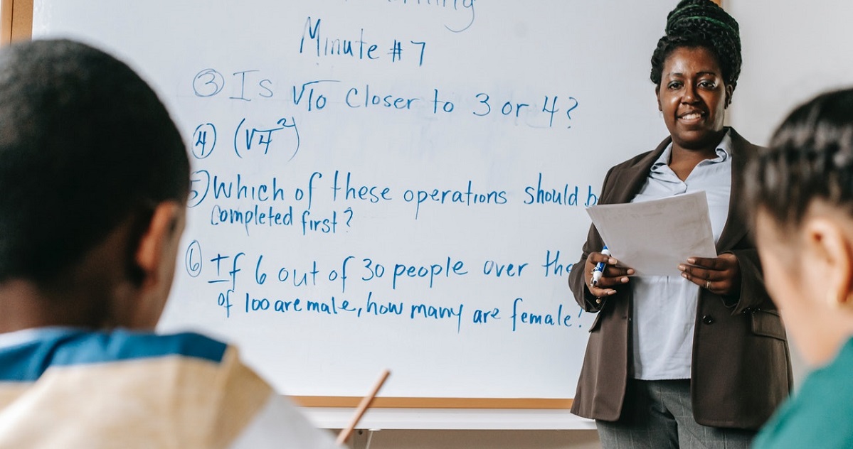 Math teacher standing in front of whiteboard in classroom; two students in foreground