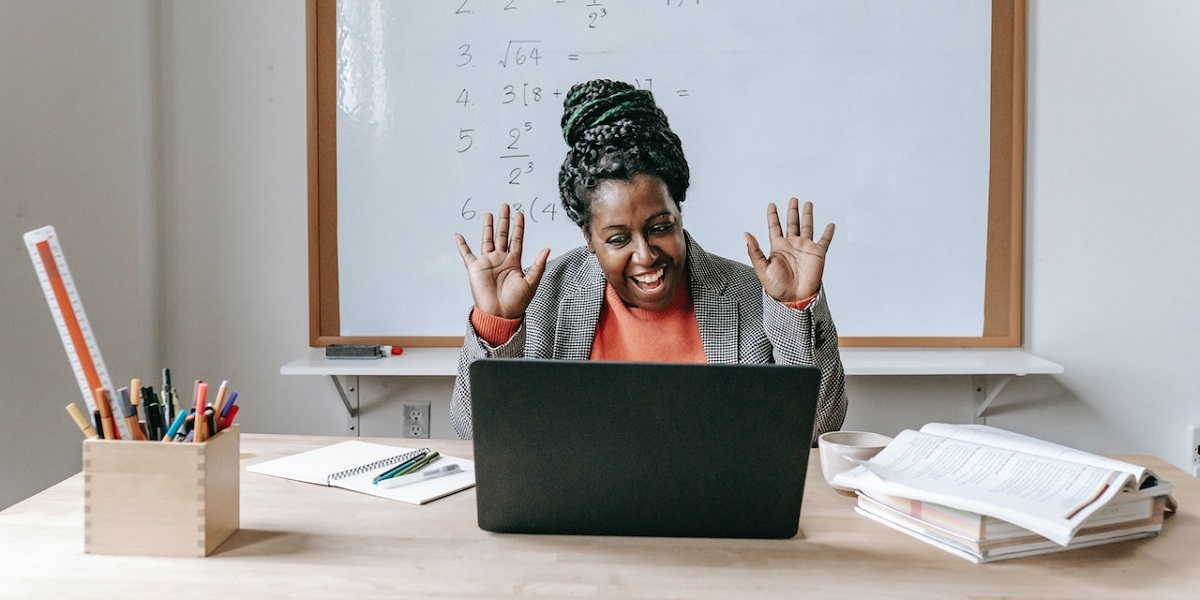 Teacher happily raising hands in front of a laptop, with whiteboard behind her that's filled with math equations