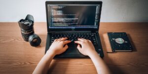 Hands typing on a laptop with a camera lens nearby on a table