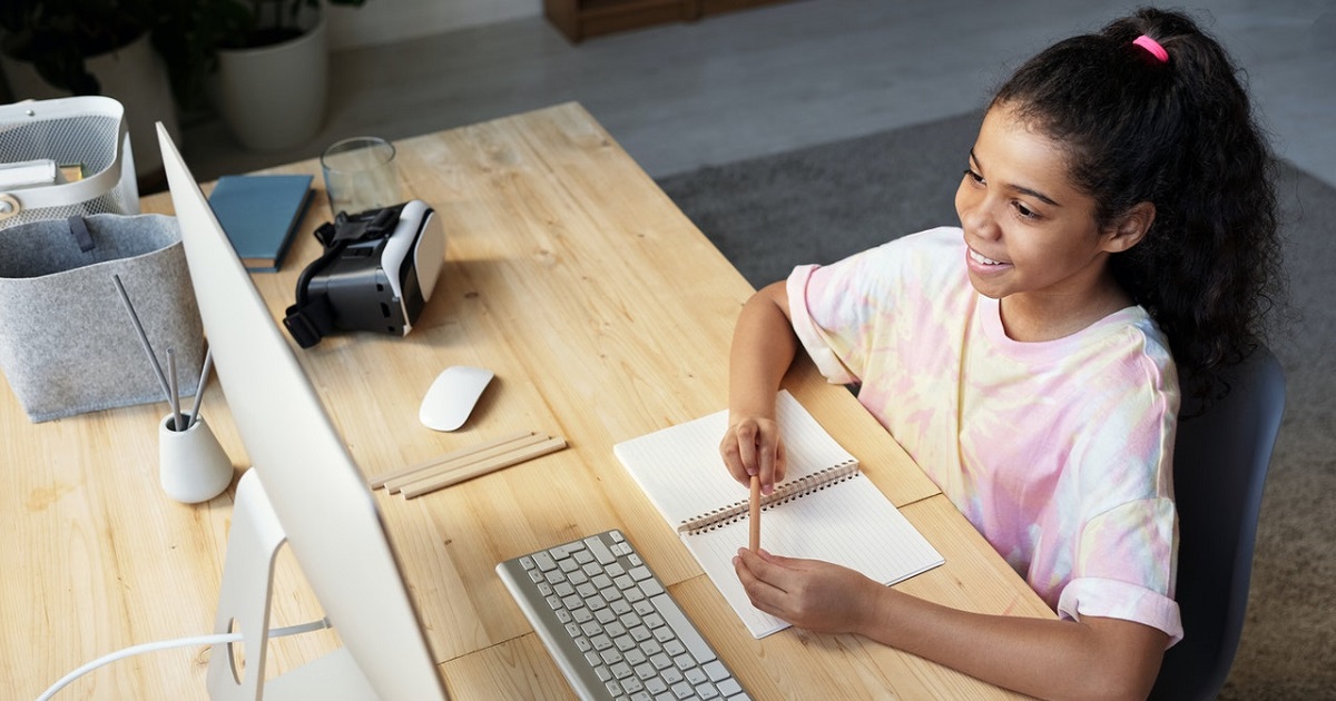 Girl looking at desktop monitor while writing in notepad on her desk