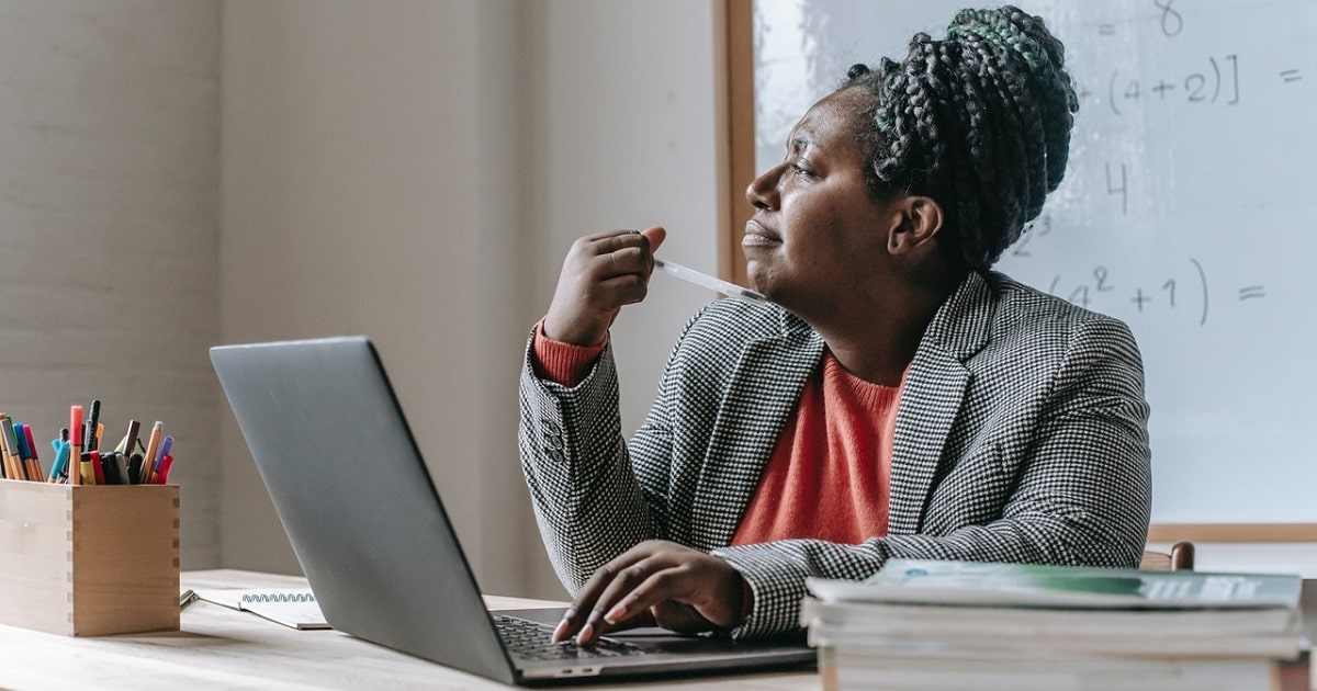 Lady staring at laptop while sitting in classroom desk; books piled on top.